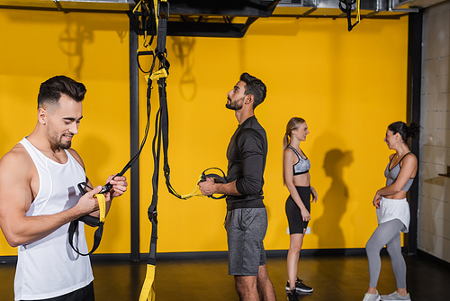 Smiling sportsman holding suspension straps near blurred multiethnic friends in gym