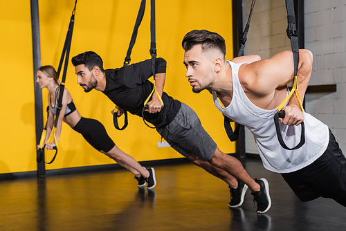 Young sportsman pulling suspension straps near blurred multiethnic friends in gym