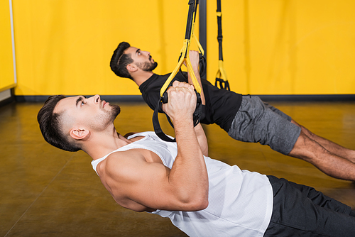 Side view of athletic sportsman pulling suspension straps near blurred arabian friend training in gym