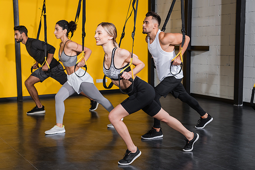 Positive sportswoman pulling suspension straps near multiethnic people in gym