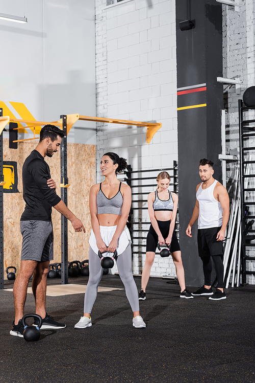 Smiling middle east sportswoman holding kettlebell near arabian trainer and people in gym