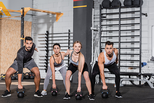 Blonde sportswoman smiling at camera near interracial friends training with kettlebells in gym