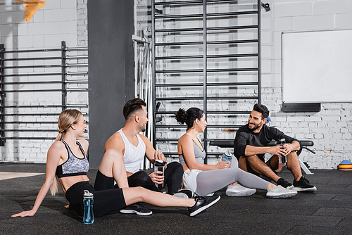 Smiling muslim man holding sports bottle near friend on floor in gym