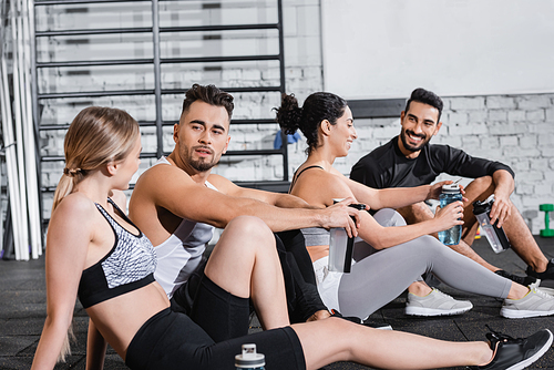 Sportsman holding sports bottle and talking to friend near interracial people in gym