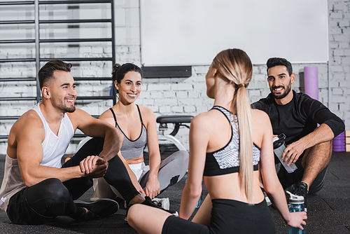 Positive multiethnic people in sportswear looking at friend near sports bottles in gym