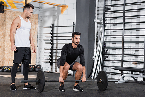 Arabian sportsman training with barbell near trainer in gym