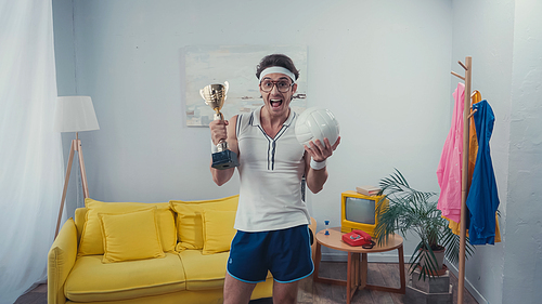 happy man holding trophy cup and sports ball while standing at home