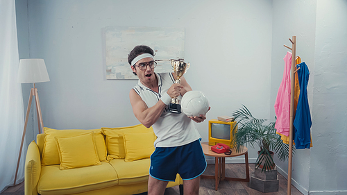excited sportsman holding trophy cup and sports ball while standing at home