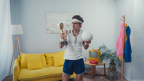 Sportsman holding trophy cup and sports ball while standing at home