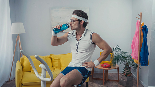 sportsman working out on exercise bike and drinking water in living room, retro sport concept