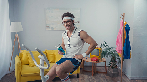 happy sportsman training on exercise bike and holding sports bottle in living room, retro sport concept