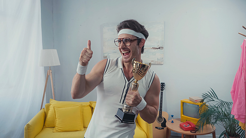 Sportsman in eyeglasses showing thumb up and holding golden trophy at home