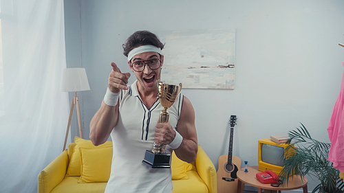 Sportsman in eyeglasses pointing with finger and holding golden trophy at home