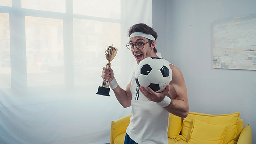Excited football player with trophy cup and soccer ball celebrating win at home