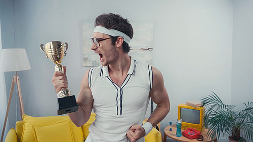 excited sportsman in eyeglasses screaming while holding golden trophy at home