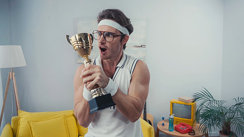 shocked sportsman in eyeglasses holding golden trophy at home