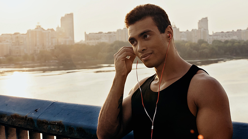 athletic bi-racial man adjusting earphones while standing on bridge over river