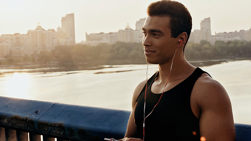 smiling bi-racial sportsman in earphones listening music on city bridge over river
