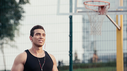young mixed race sportsman in earphones looking away near blurred basketball ring