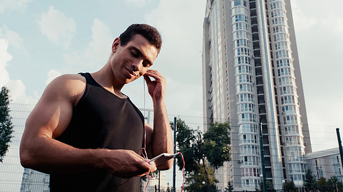 smiling bi-racial sportsman with smartphone inserting earphone outdoors