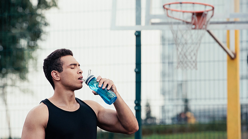 mixed race sportsman with closed eyes drinking water from sports bottle outdoors