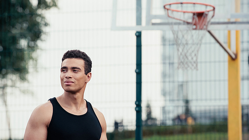 athletic bi-racial man looking away near blurred basketball ring outdoors