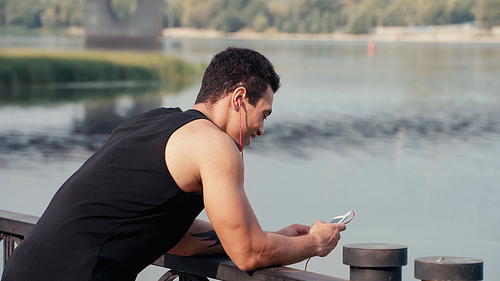 happy and sportive bi-racial man in earphones using smartphone on bridge over river