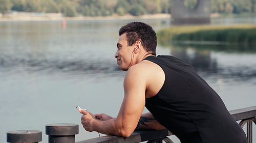smiling bi-racial man with smartphone listening music on bridge over river