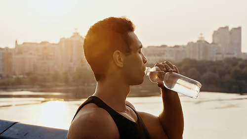 athletic mixed race man drinking water over river in city