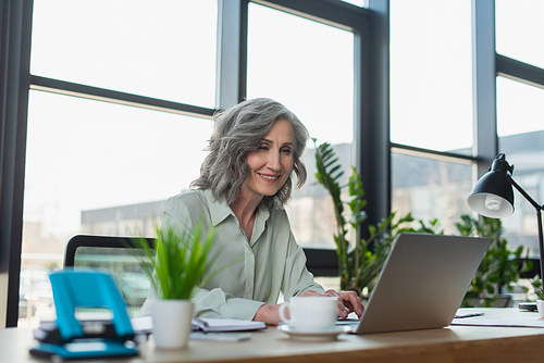 Smiling businesswoman using laptop near coffee and stationery in office