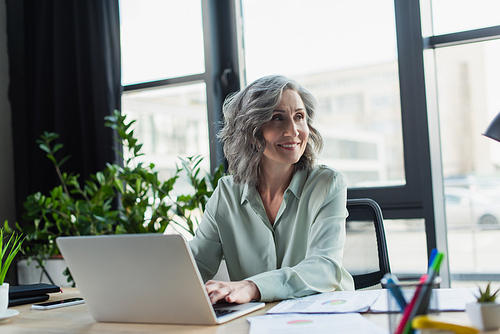 Smiling mature businesswoman using laptop near papers in office