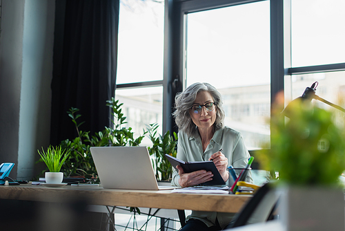 Mature businesswoman in eyeglasses writing on notebook near laptop and plant in office