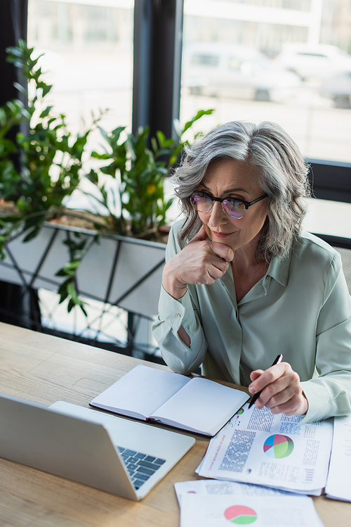Grey haired businesswoman holding pen near paperwork, notebook and laptop in office