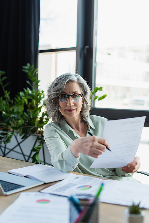 Mature businesswoman holding documents near blurred laptop and notebook in office