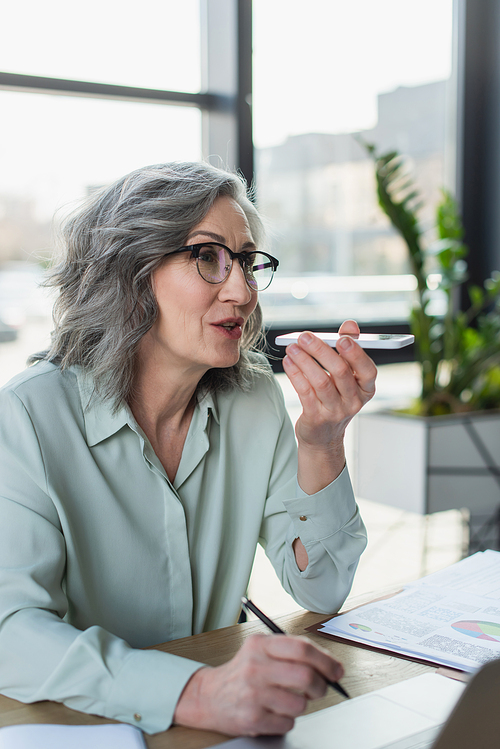 Grey haired businesswoman recording voice message on cellphone and holding pen near laptop in office