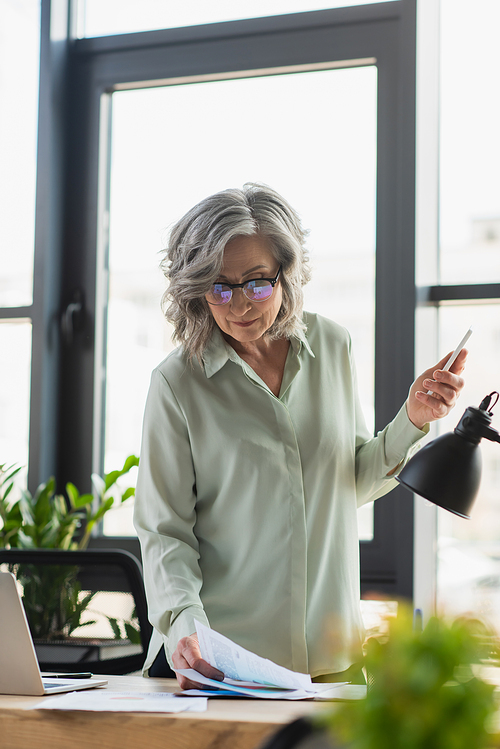 Mature businesswoman in eyeglasses holding smartphone near documents in office