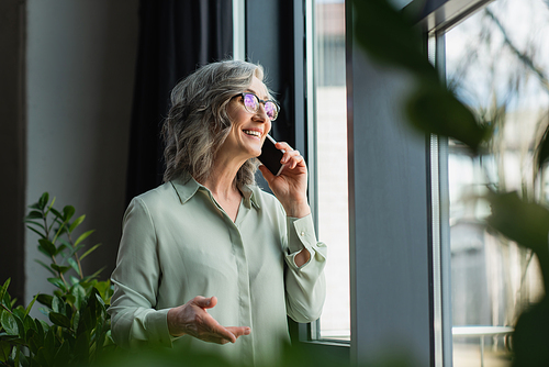 Positive businesswoman talking on smartphone near plants in office