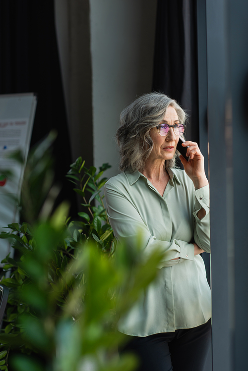 Mature businesswoman talking on mobile phone near blurred plants in office