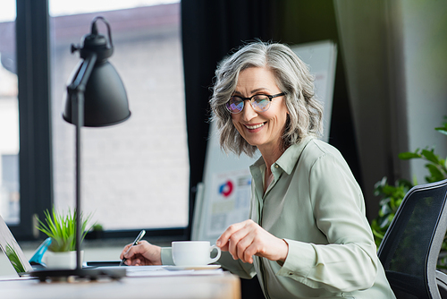 Happy businesswoman in eyeglasses taking coffee cup near laptop and papers