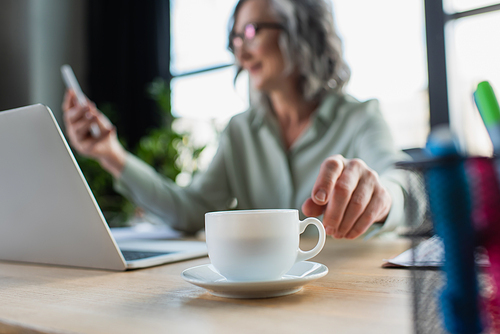 Cup of coffee near blurred businesswoman and laptop in office