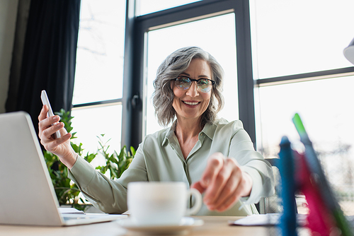 Happy businesswoman holding smartphone and taking cup of coffee in office