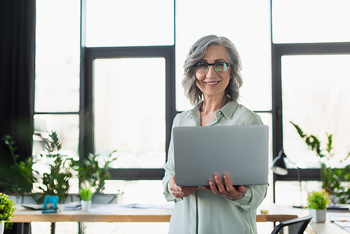 Cheerful grey haired businesswoman in eyeglasses holding laptop and  in office