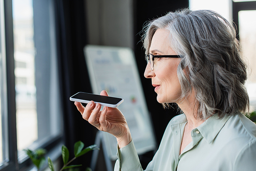 Side view of mature businesswoman in eyeglasses recording voice message on smartphone in office