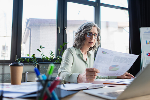 Grey haired businesswoman holding paper near coffee to go and laptop in office
