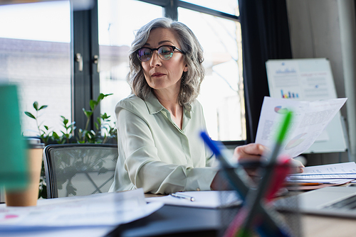 Grey haired businesswoman in eyeglasses looking at papers near laptop and takeaway drink in office
