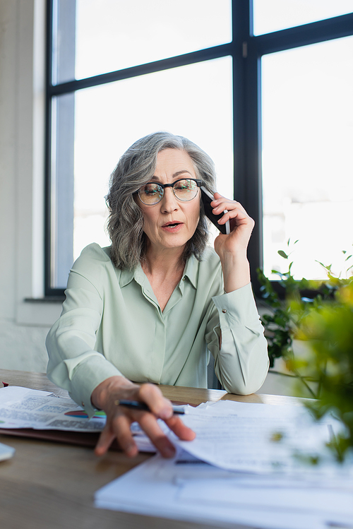 Businesswoman talking on cellphone near blurred papers on table in office