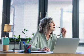 Pensive businesswoman holding eyeglasses near laptop in office