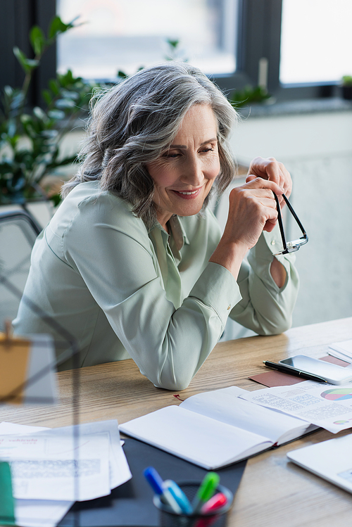 Cheerful businesswoman holding eyeglasses near documents and gadgets on table in office