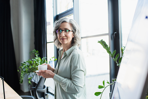 Grey haired businesswoman holding coffee near flip chart in office