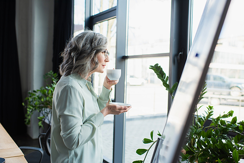 Side view of mature businesswoman holding cup of coffee near blurred flip chart in office